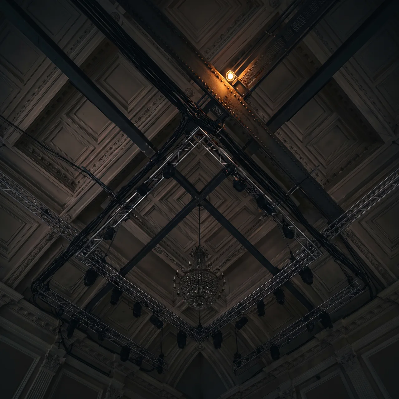 Looking straight up at a coffered ceiling with lighting truss, cabling and an unlit chandelier.